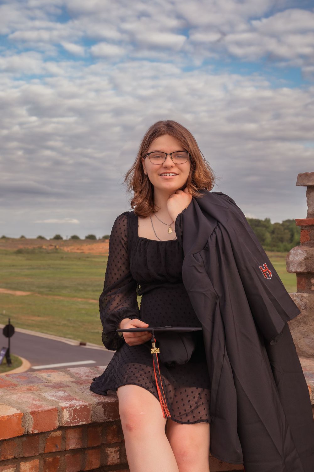 A person wearing a black dress and graduation gown poses on a brick wall with a cloudy sky background.