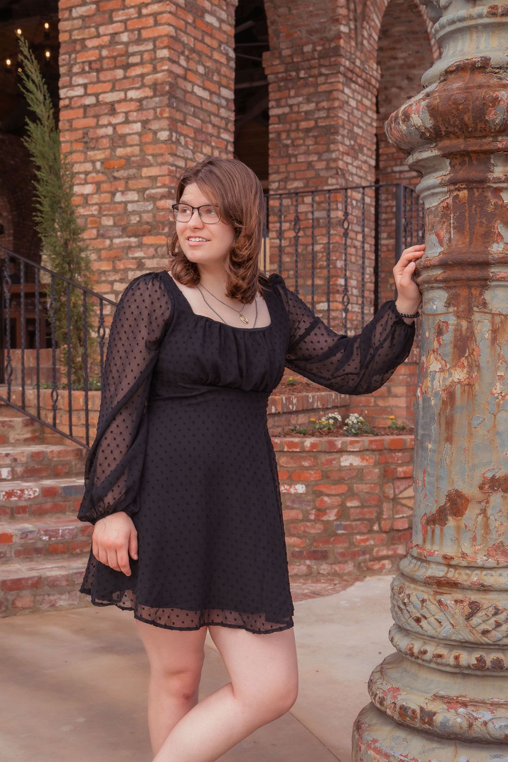 Elegant black lace dress with long sleeves photographed against vintage brick columns and decorative stone pillars.