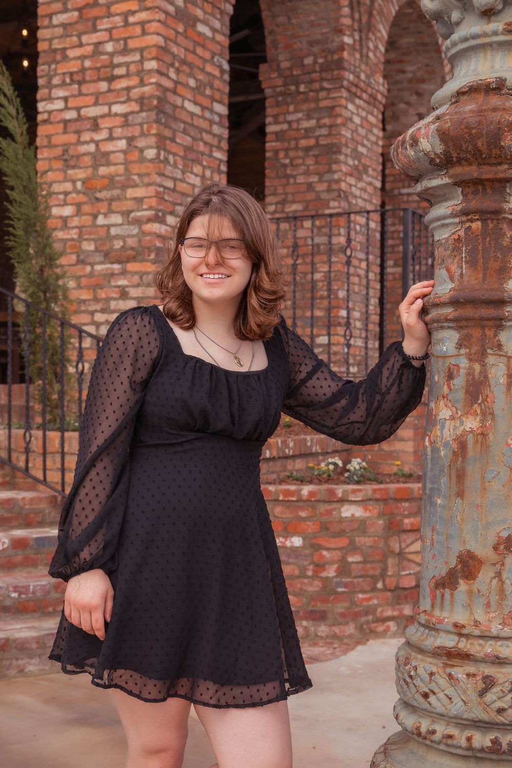 A person in a black polka dot babydoll dress poses near red brick arches and stone columns.
