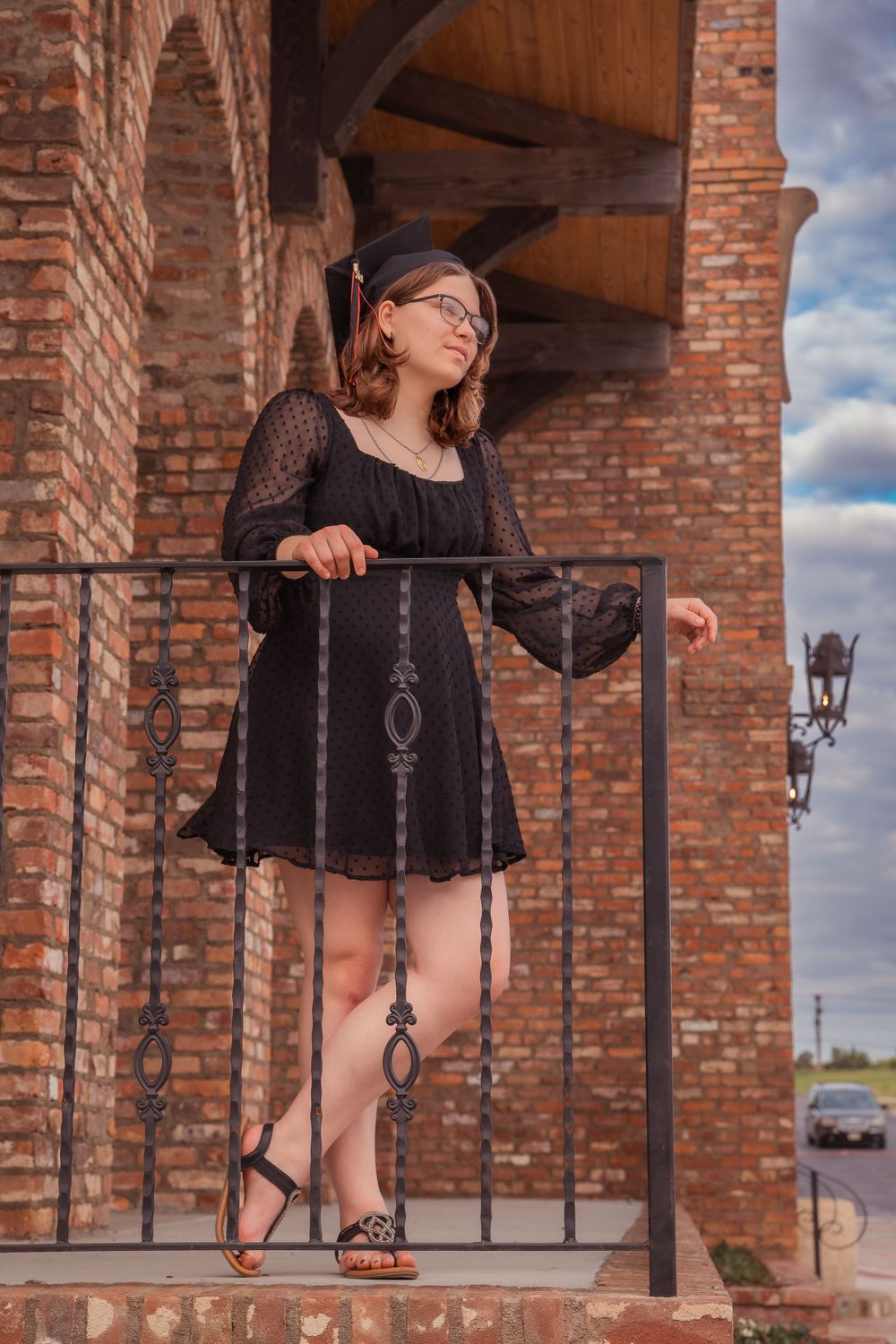 A person in a black dress poses on a brick archway balcony against a cloudy sky.