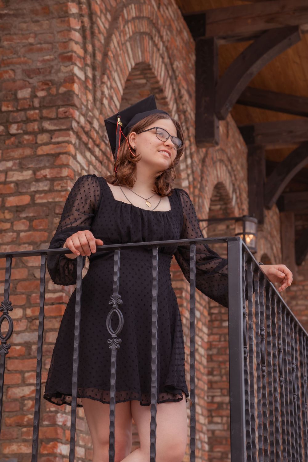 Graduate in black lace dress standing on brick building balcony with ornate metal railing.