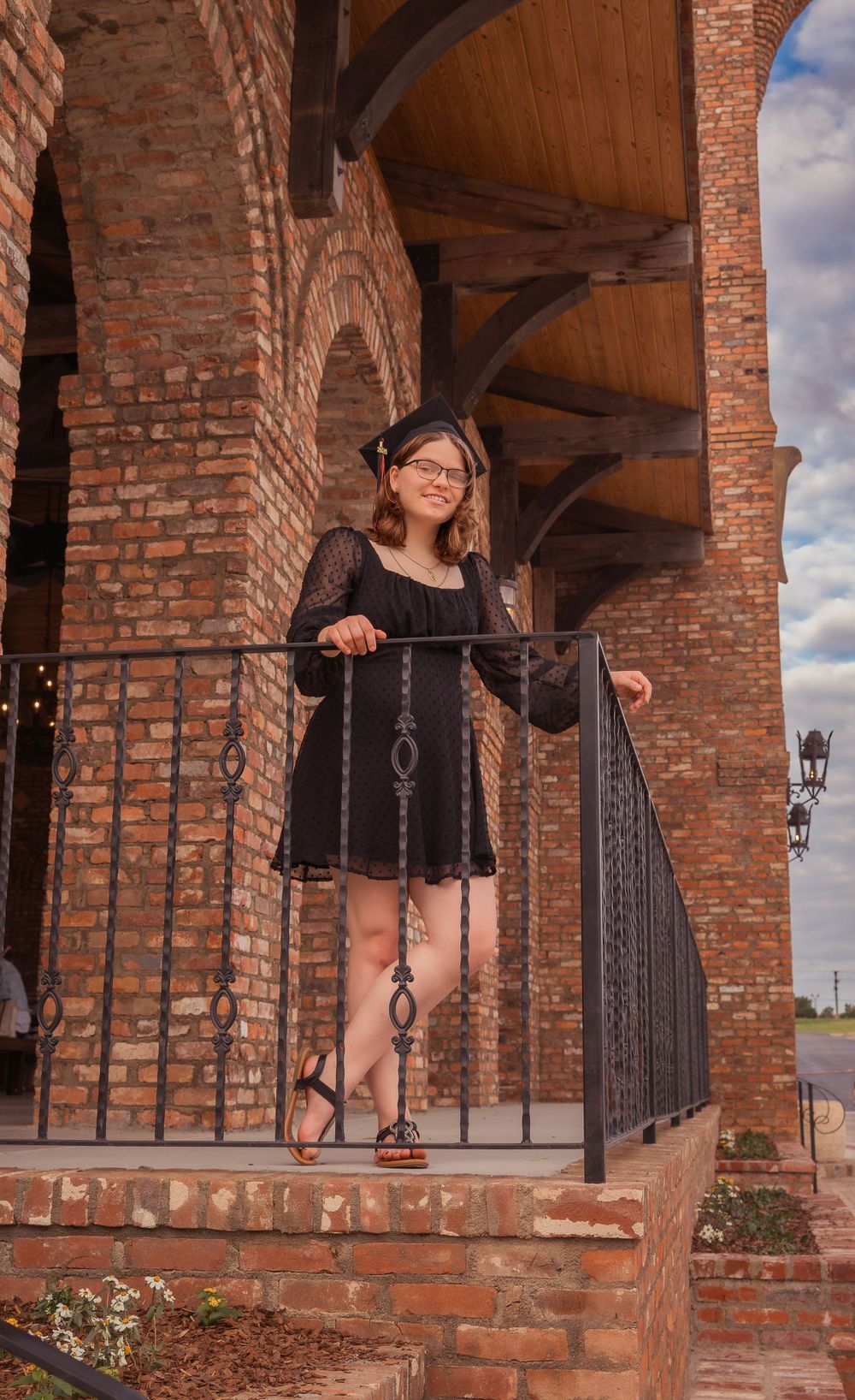 A person stands on brick steps in a black summer dress, leaning against an iron railing under arched brick architecture.