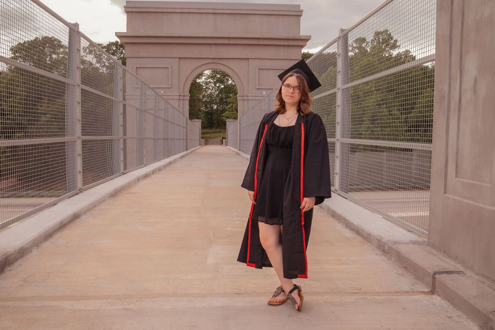 Graduate stands on covered outdoor walkway in black and red graduation regalia with elegant decor.