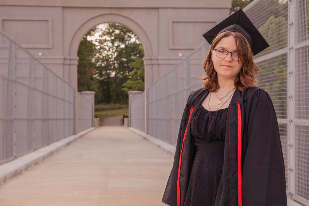 Graduate in cap and gown stands on a covered walkway with archways and greenery visible in background.