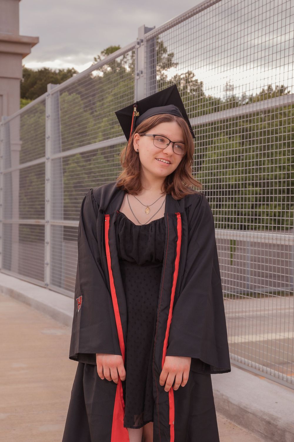 A graduate in black academic regalia with red trim stands smiling beside a mesh fence on graduation day.