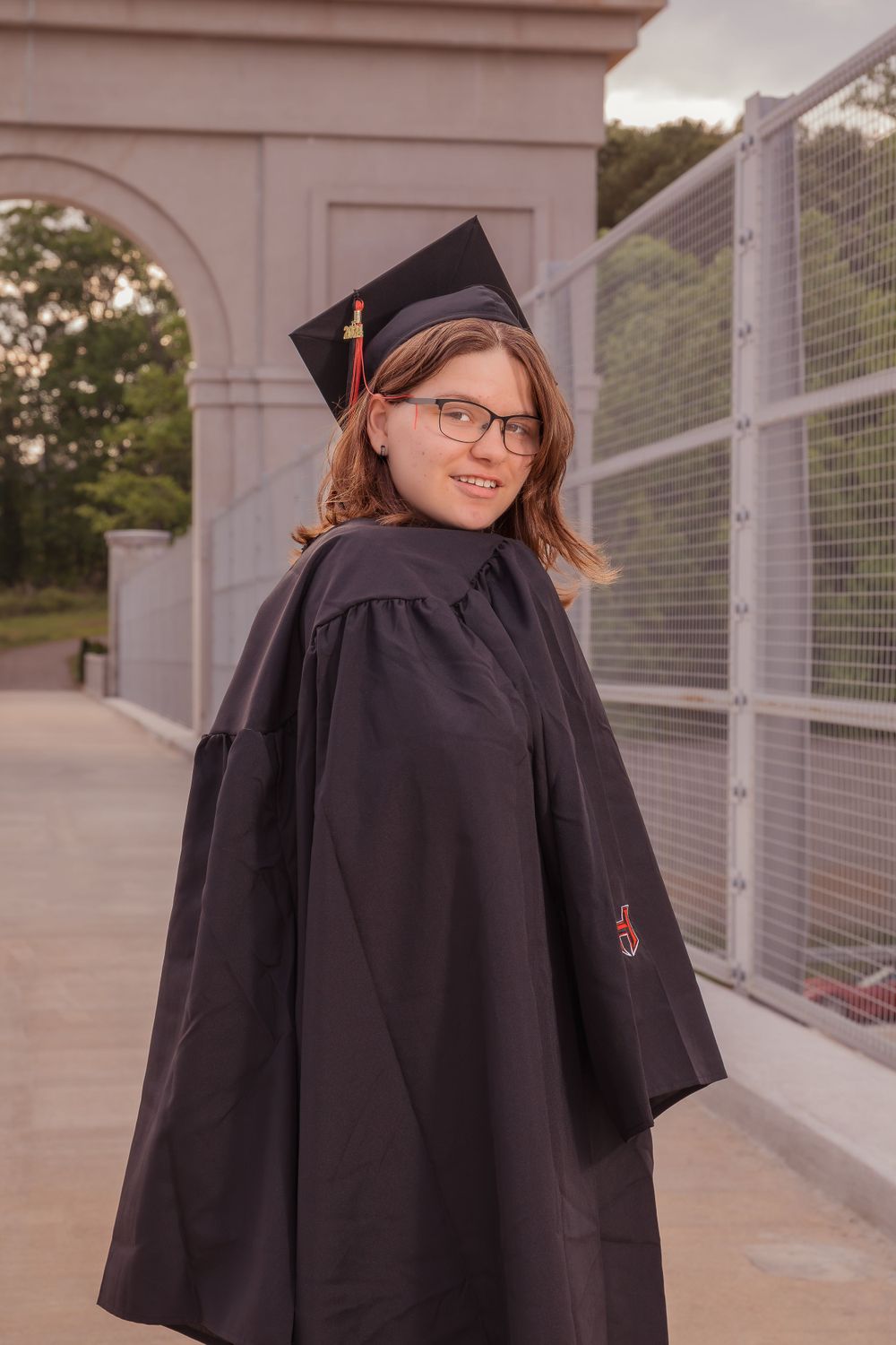 Graduate in black cap and gown stands outdoors near archway in golden evening light wearing glasses.