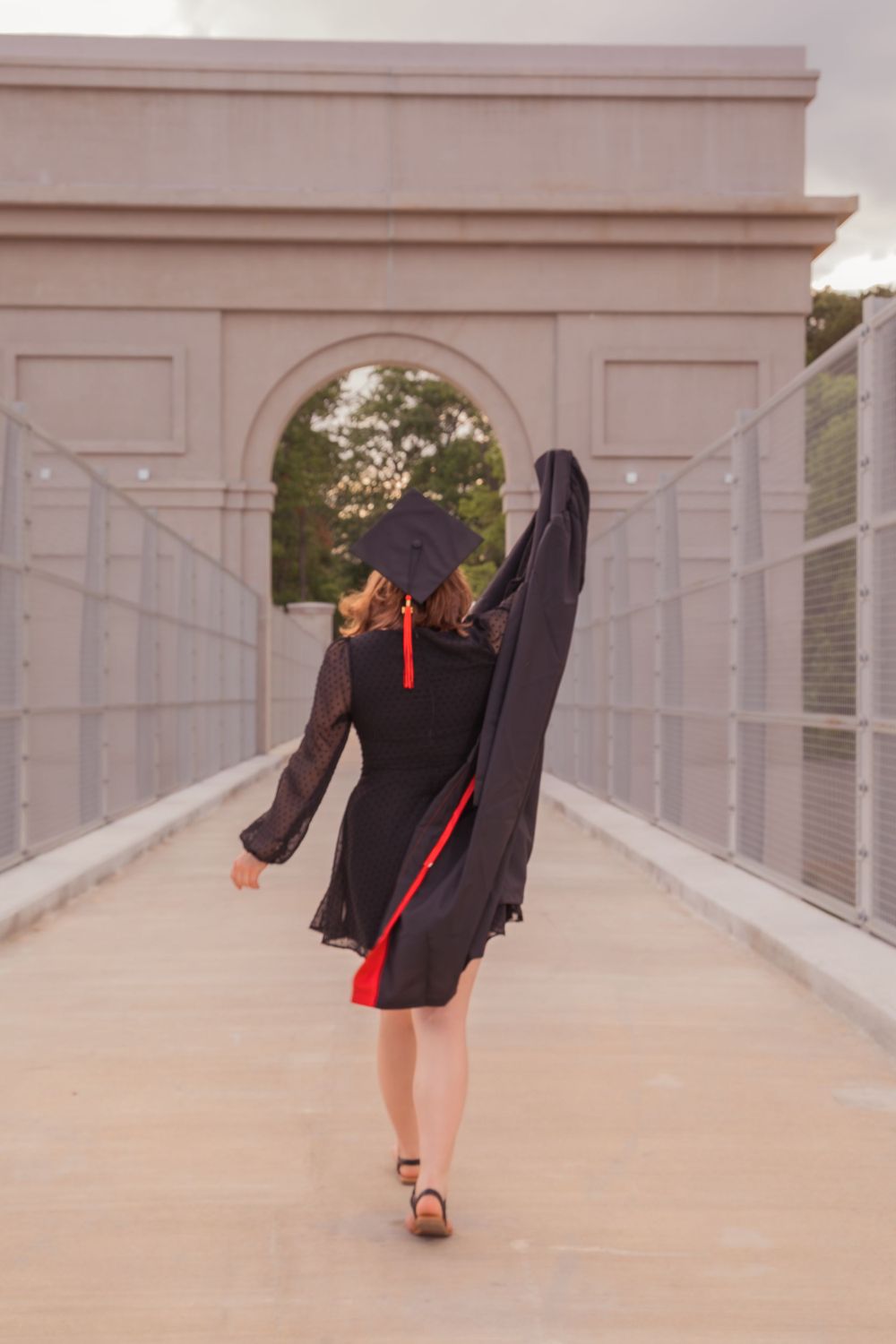 Graduate in black cap and gown twirls joyfully on a white bridge with an arched architectural backdrop at sunset.