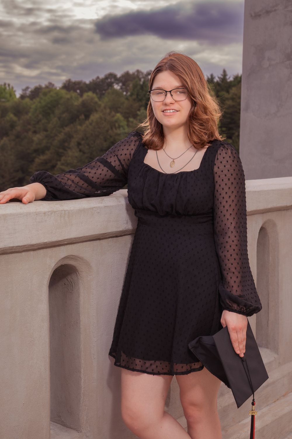 Someone poses on a balcony wearing a black polka dot dress with sheer sleeves and holding a clutch purse against moody skies.