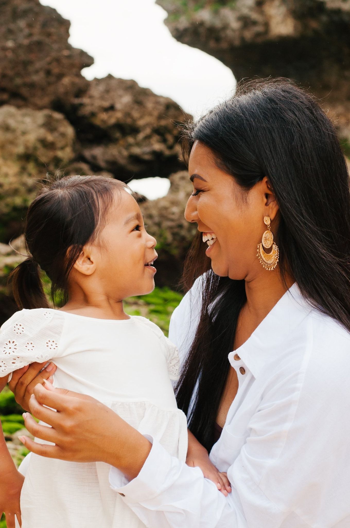 Mother and daughter share joyful moment while wearing white against rocky backdrop.