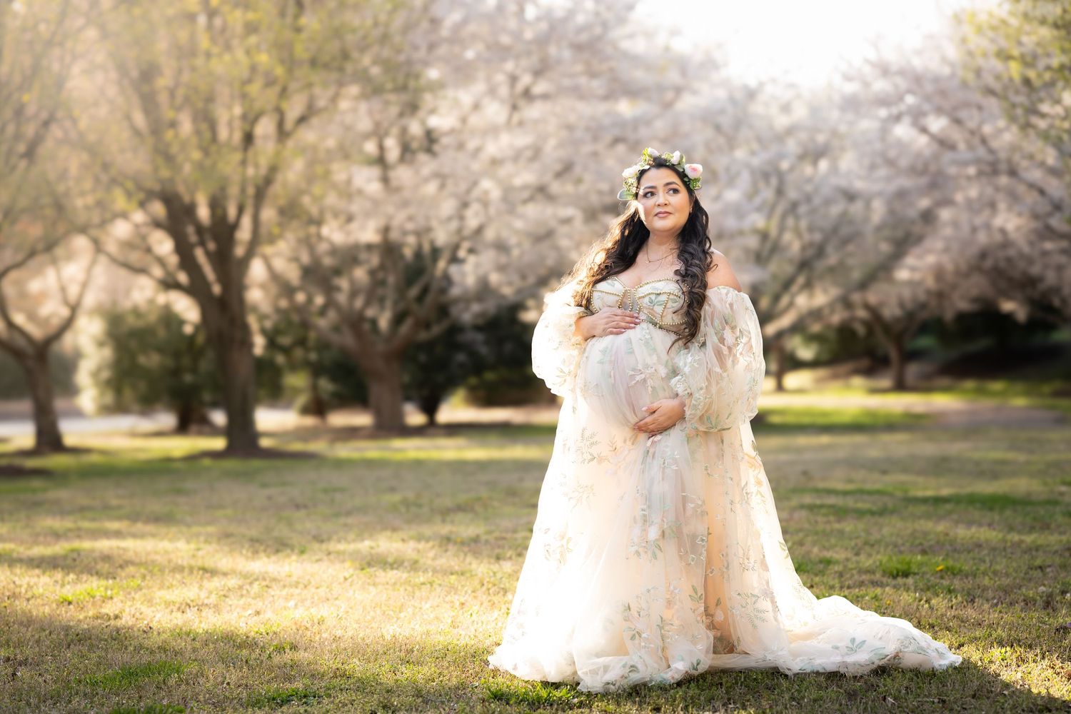 A bride in a lace wedding dress poses outdoors among blooming cherry blossom trees during golden hour.