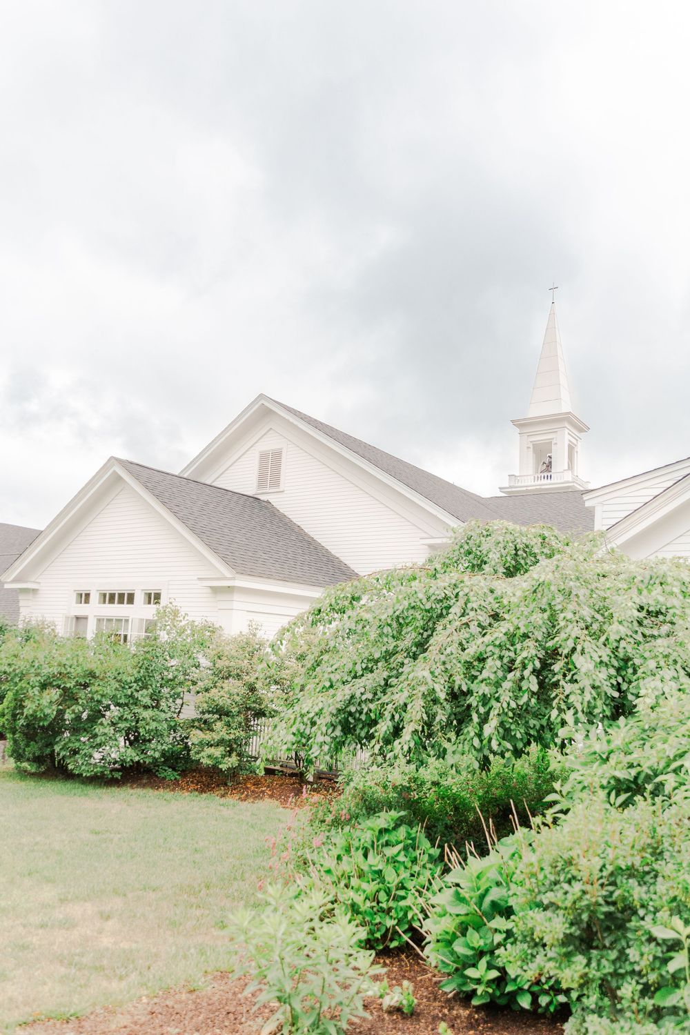 White church building with steeple surrounded by lush green bushes under cloudy sky.