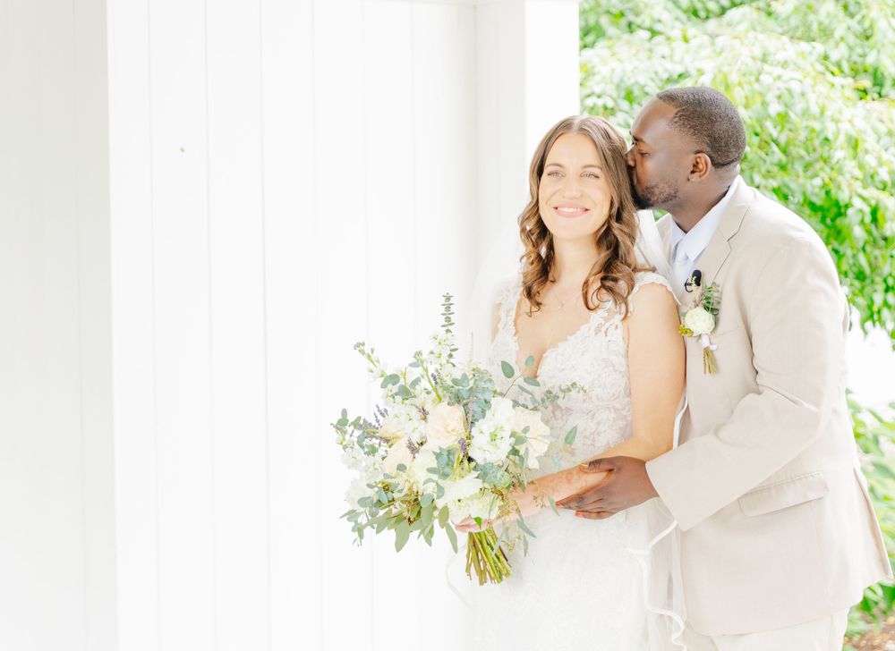 Wedding couple shares tender moment on white porch with elegant floral bouquet.