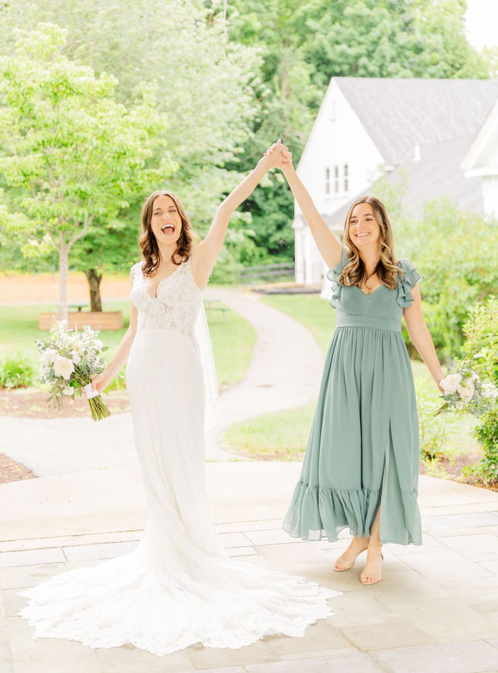 Two happy women celebrate on garden pathway with bridal bouquet and flowing dresses.