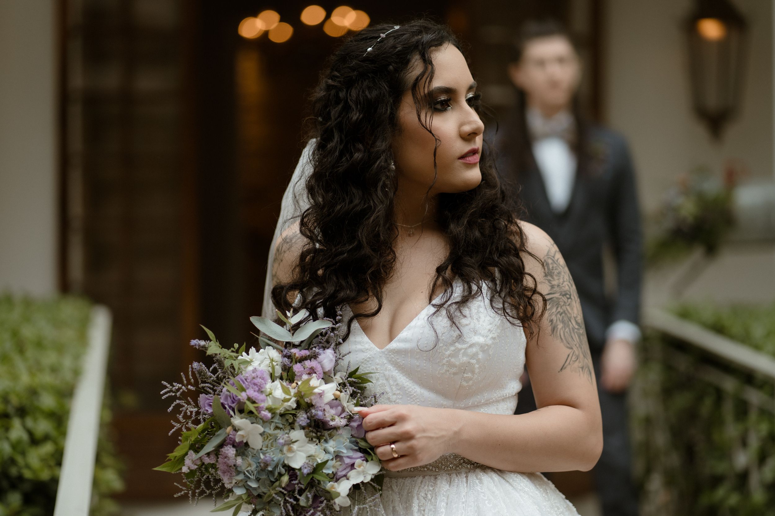 Bride holds purple wildflower bouquet in elegant white gown with lace details.