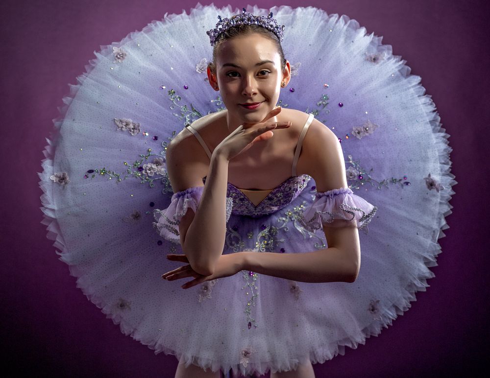 Ballerina poses in white tutu with purple accents against dark background.