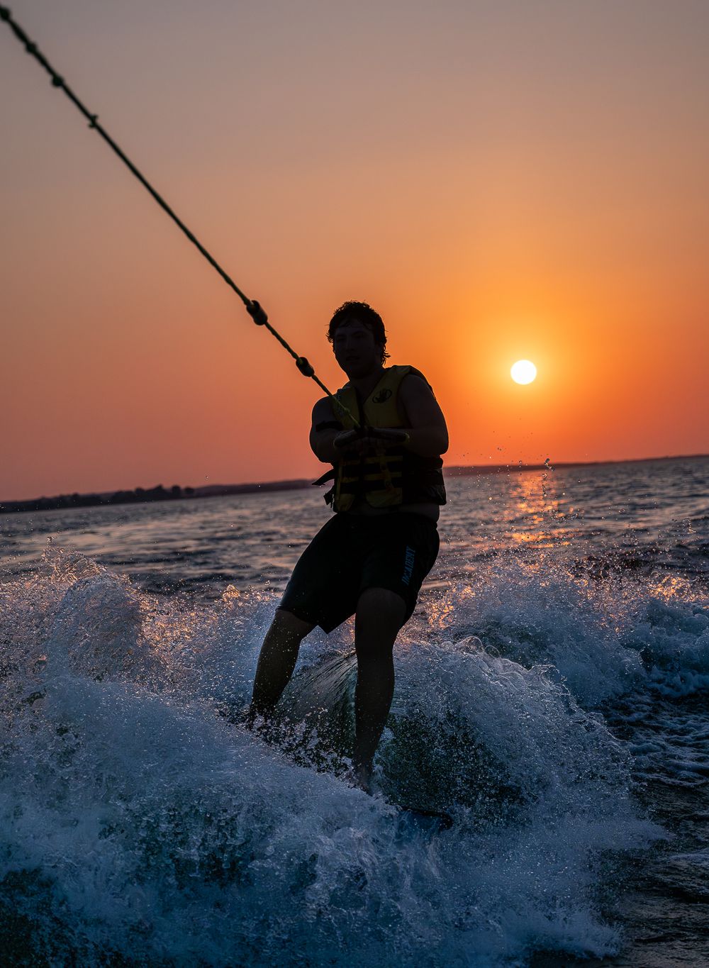 Silhouette of wake surfer against orange sunset creating dramatic splash.