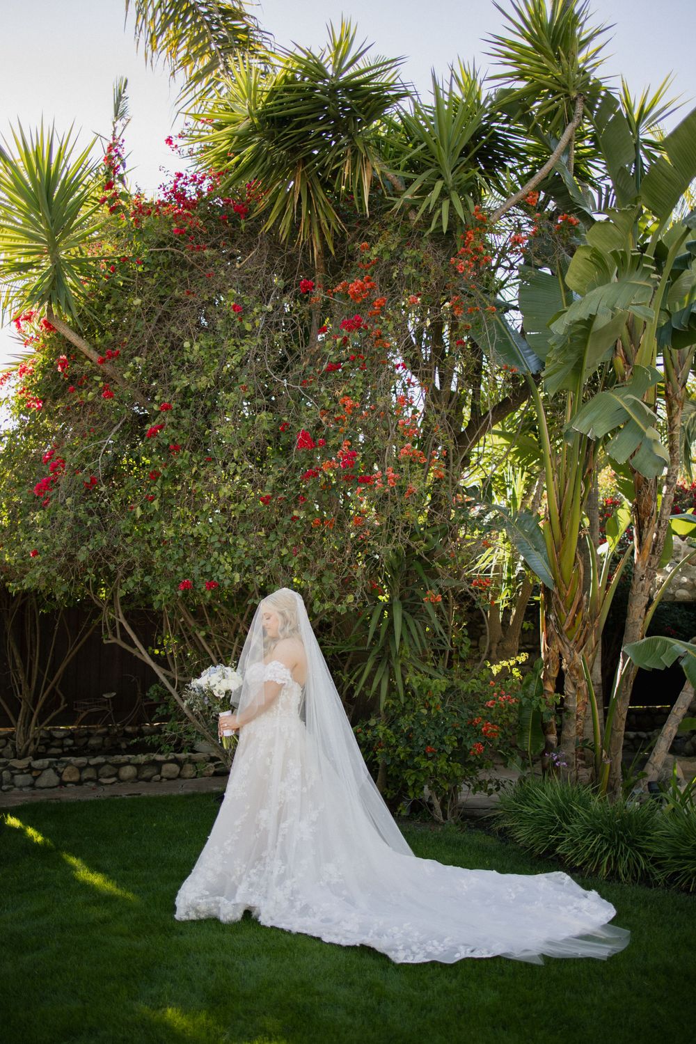A wedding dress with a flowing train and veil photographed in a lush tropical garden with palm trees and red flowers.