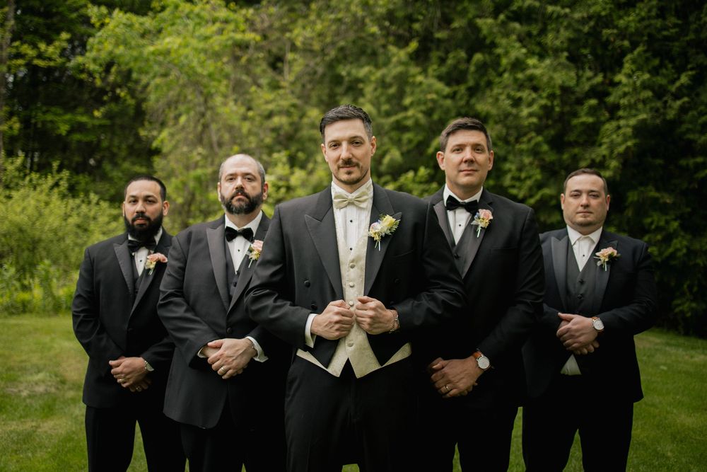 A wedding party in black tuxedos and formal attire poses together outdoors in a garden setting.