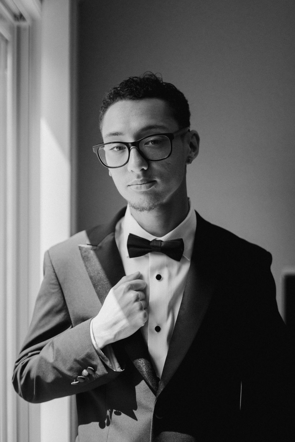 Black and white photo series showing someone adjusting their bowtie while wearing a formal tuxedo near windows.