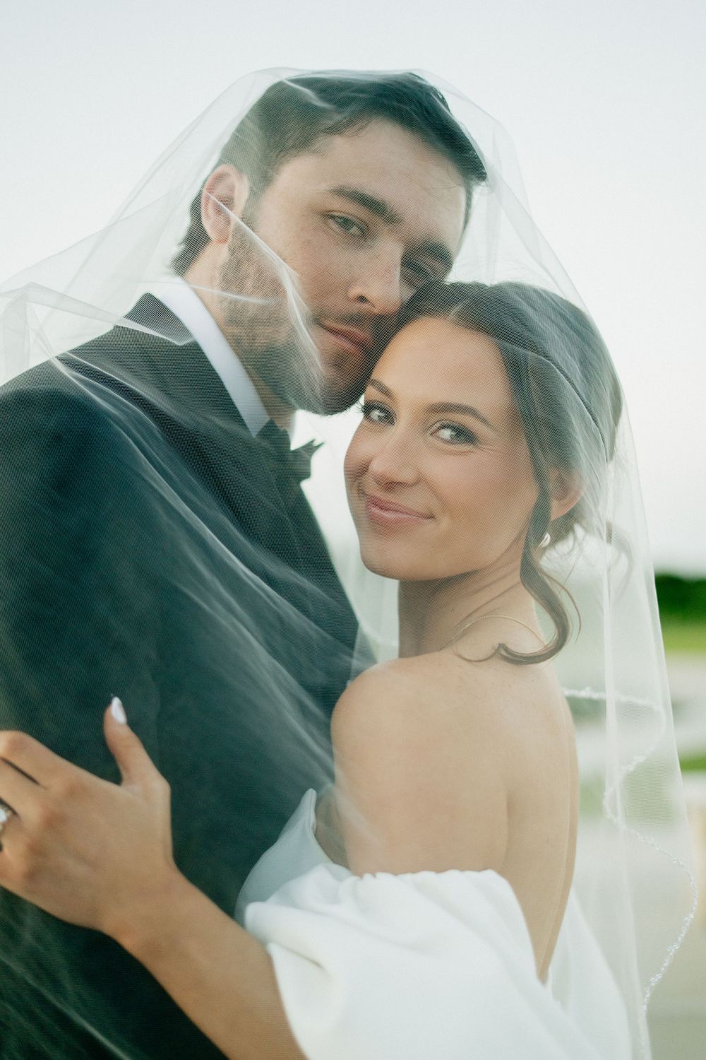 A romantic wedding portrait shows a couple embracing under a delicate veil outdoors.