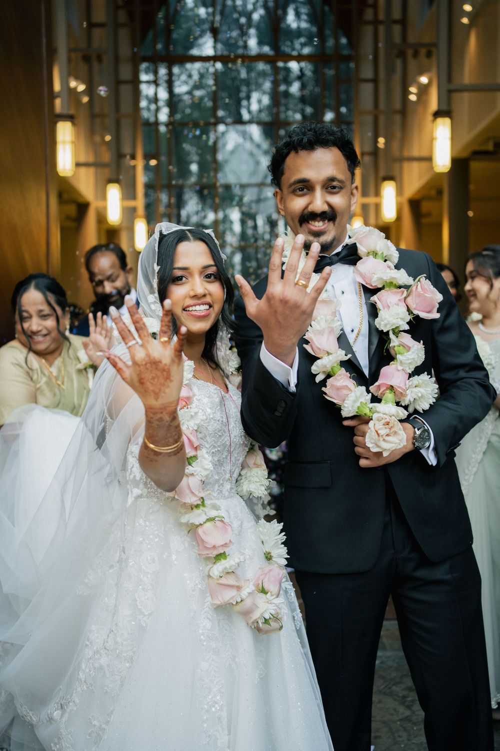 Newlyweds show off their rings with joyful smiles while holding a floral garland at an indoor wedding ceremony.