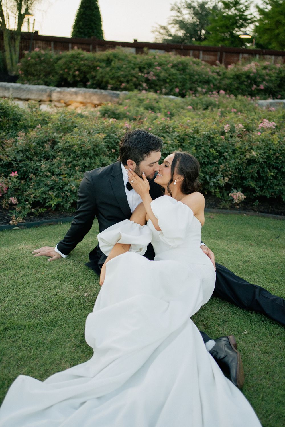 A couple in wedding attire share romantic moments on a grass lawn surrounded by blooming rose bushes in a garden setting.