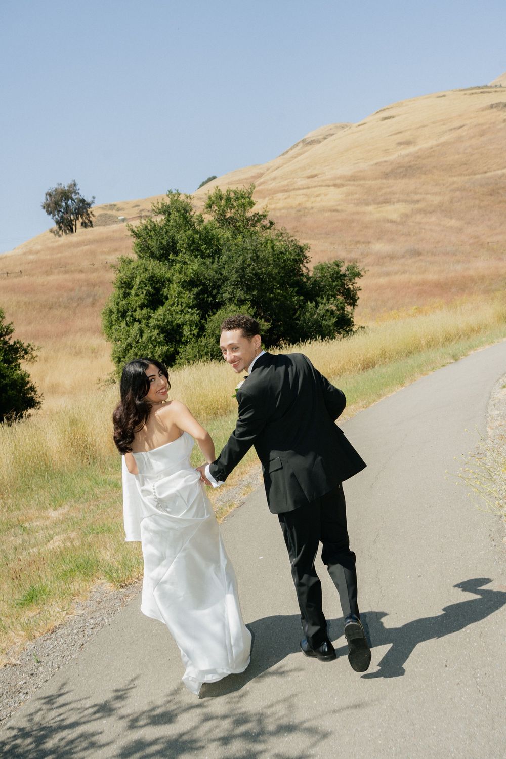 A couple in wedding attire walks hand in hand along a dirt path with golden hills in the background.