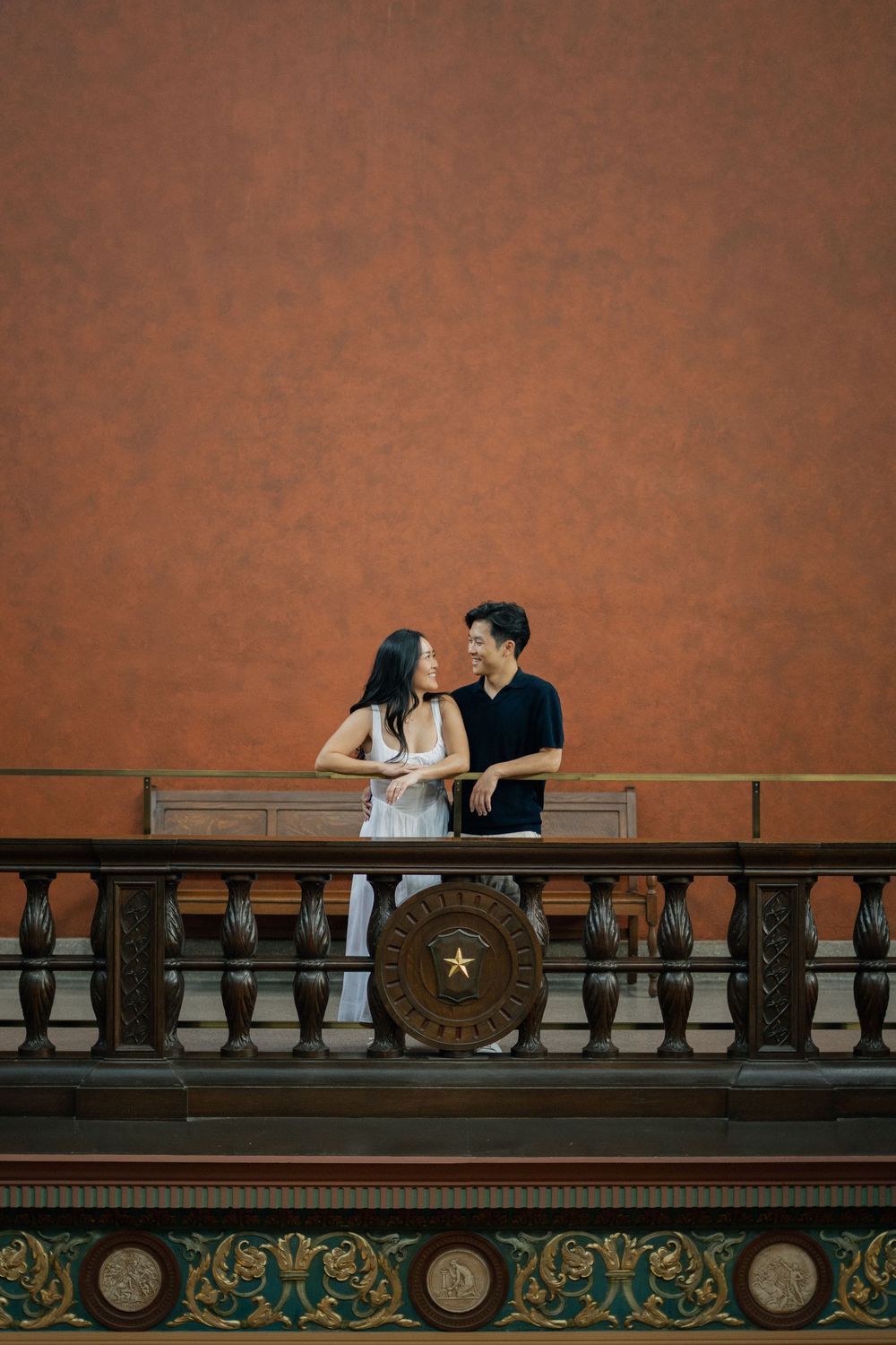 A couple shares a romantic moment against a rustic orange wall with an ornate wooden railing featuring a lone star emblem.