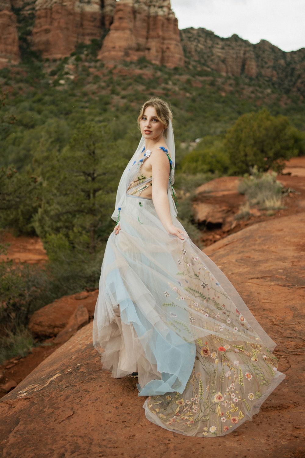 An ethereal wedding dress with floral details flows in the wind against red rock cliffs.