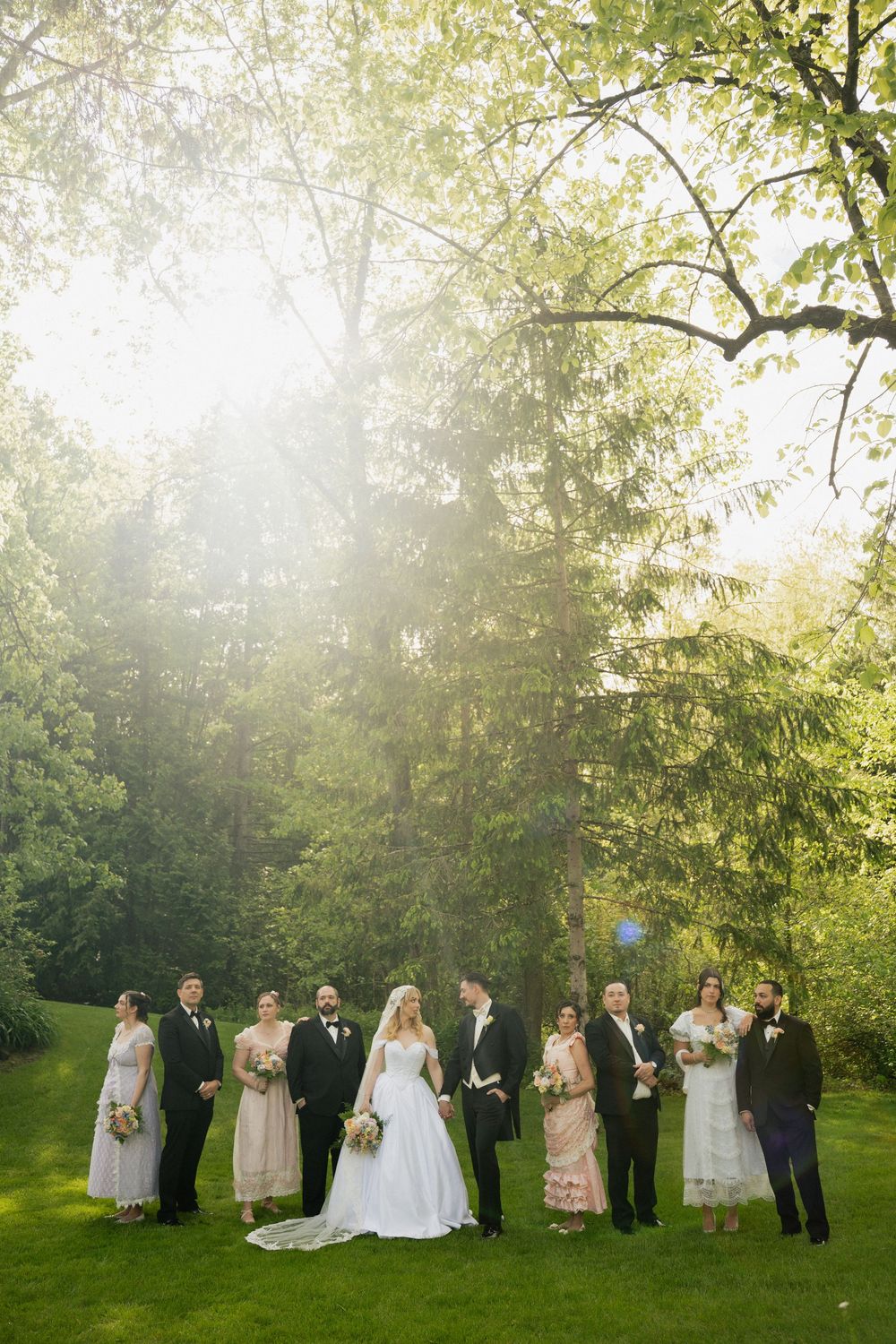 A wedding party lineup stands in front of sunlit trees on a green lawn during golden hour.
