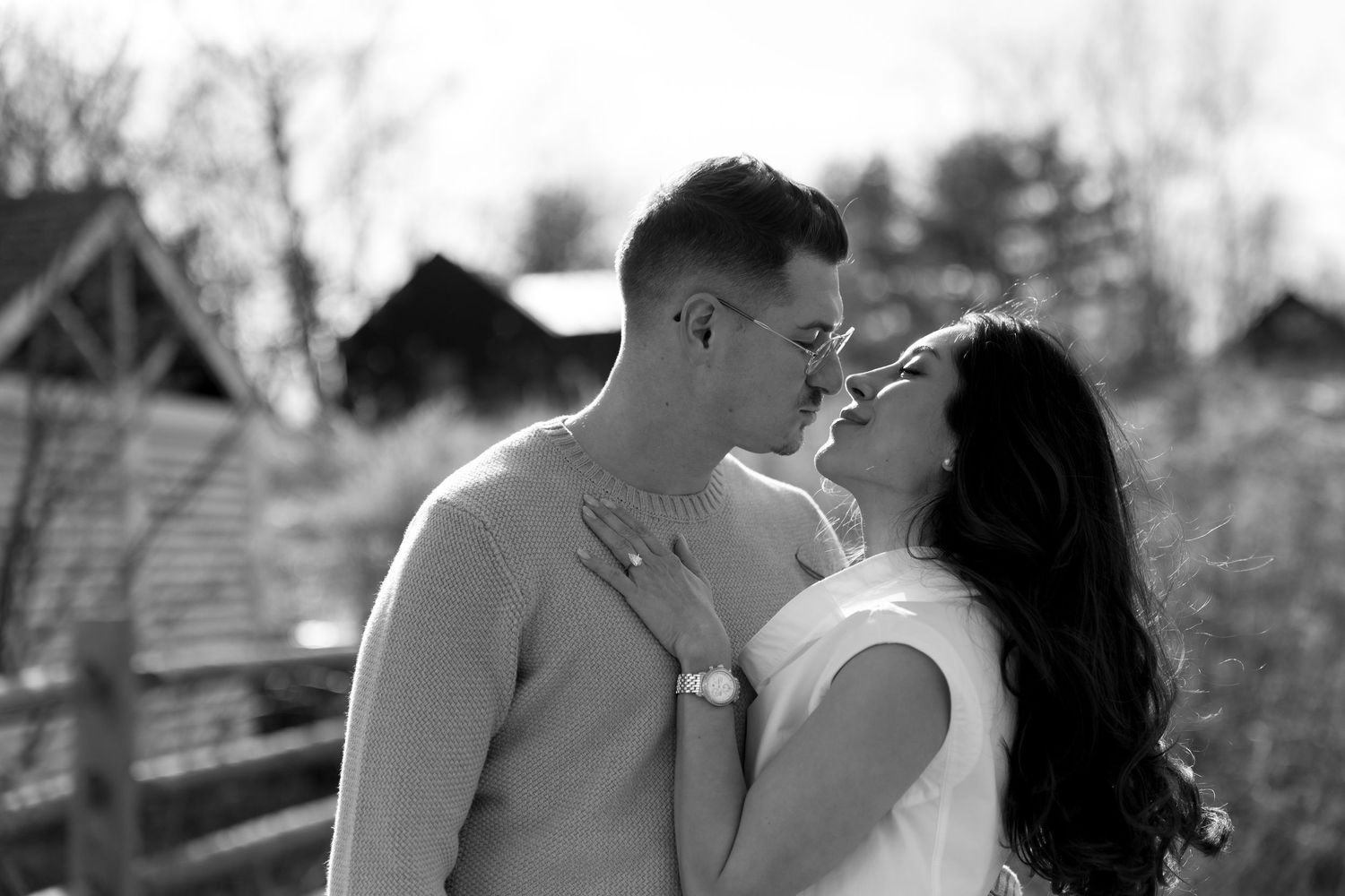 Black and white photo of a couple sharing a romantic kiss outdoors near rustic barn buildings.