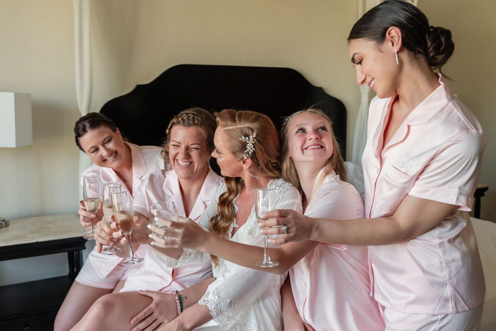 Group in matching pink robes sharing drinks and laughter during pre-wedding preparations.