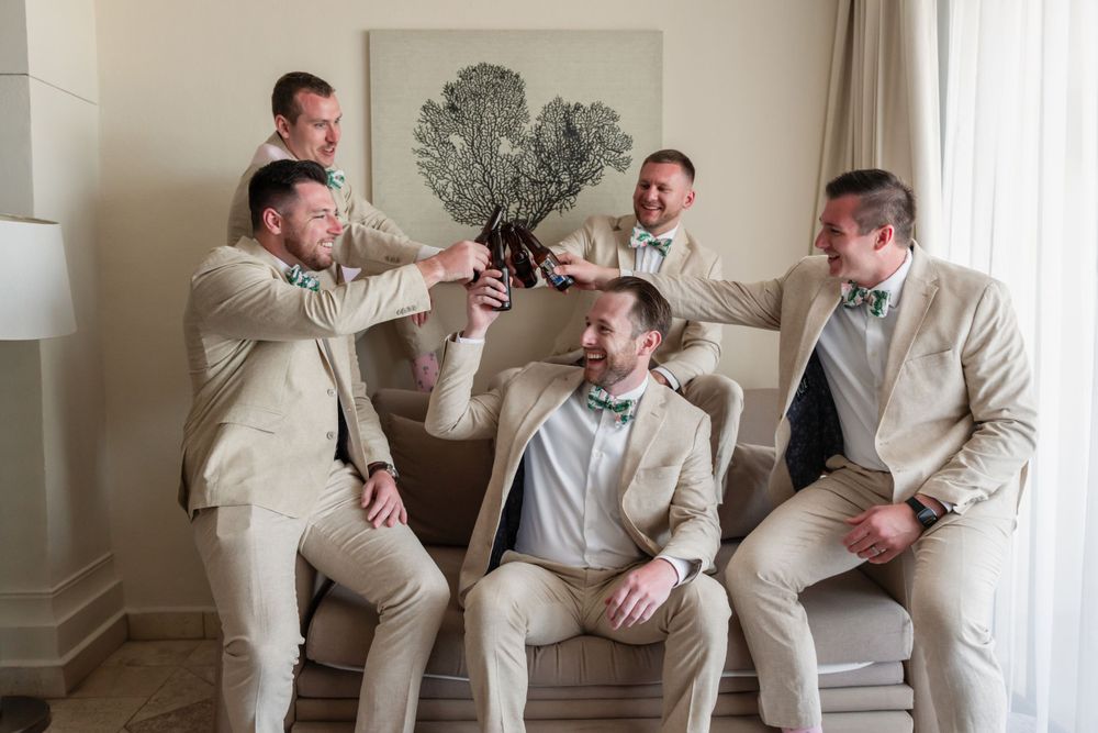 Groomsmen in beige suits toast and celebrate in a light-filled room.