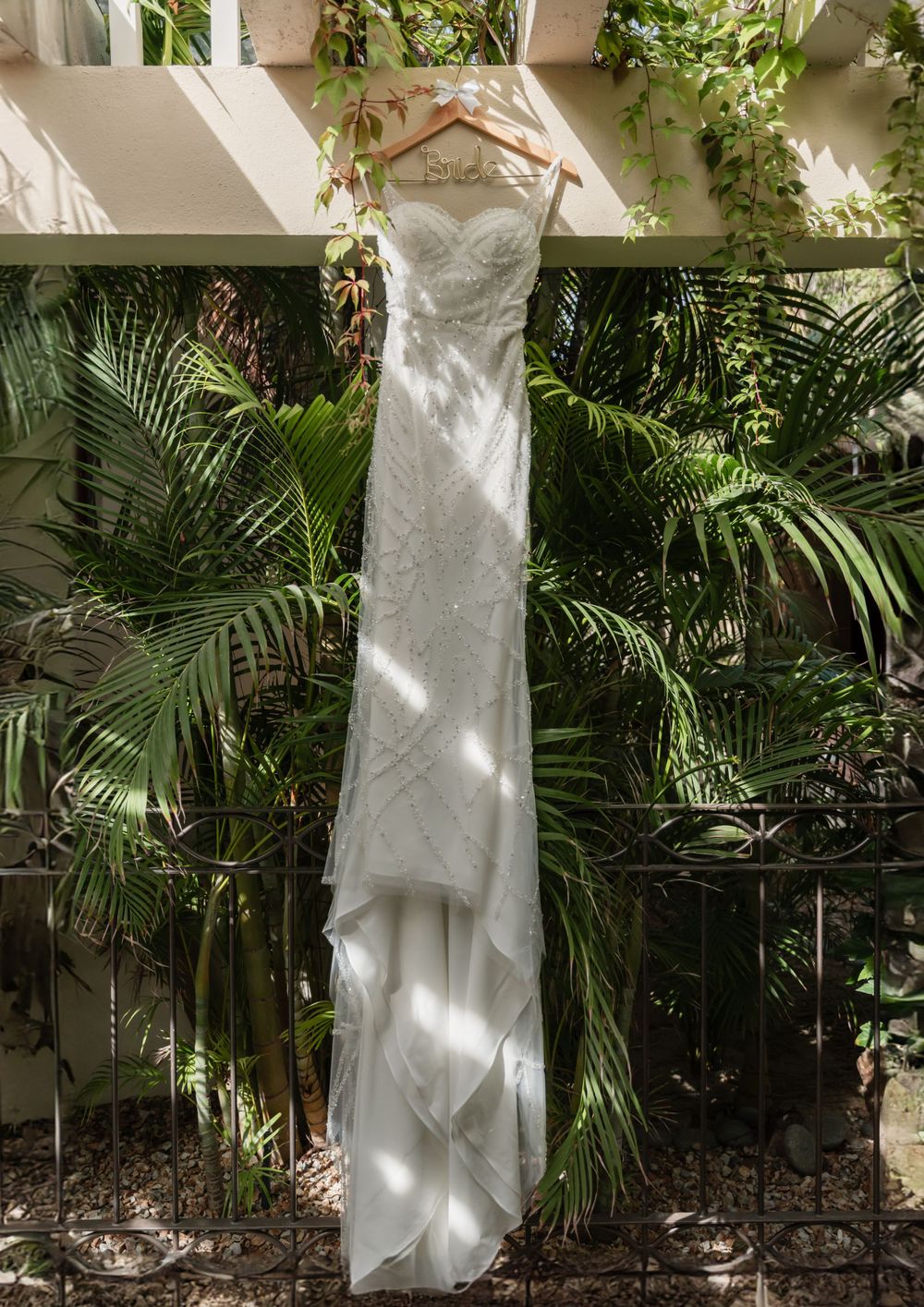 White wedding dress hanging on display against tropical foliage and palm leaves.