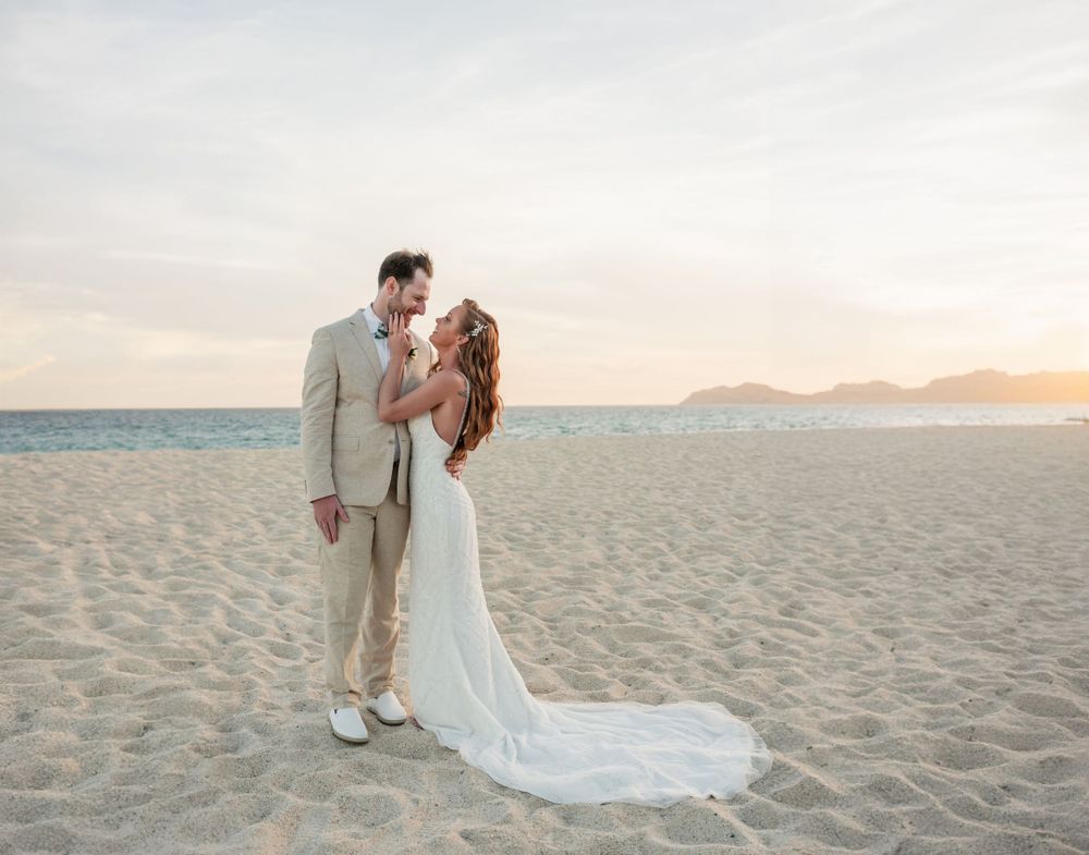 A couple shares a romantic moment on a beach at sunset in formal wedding attire.