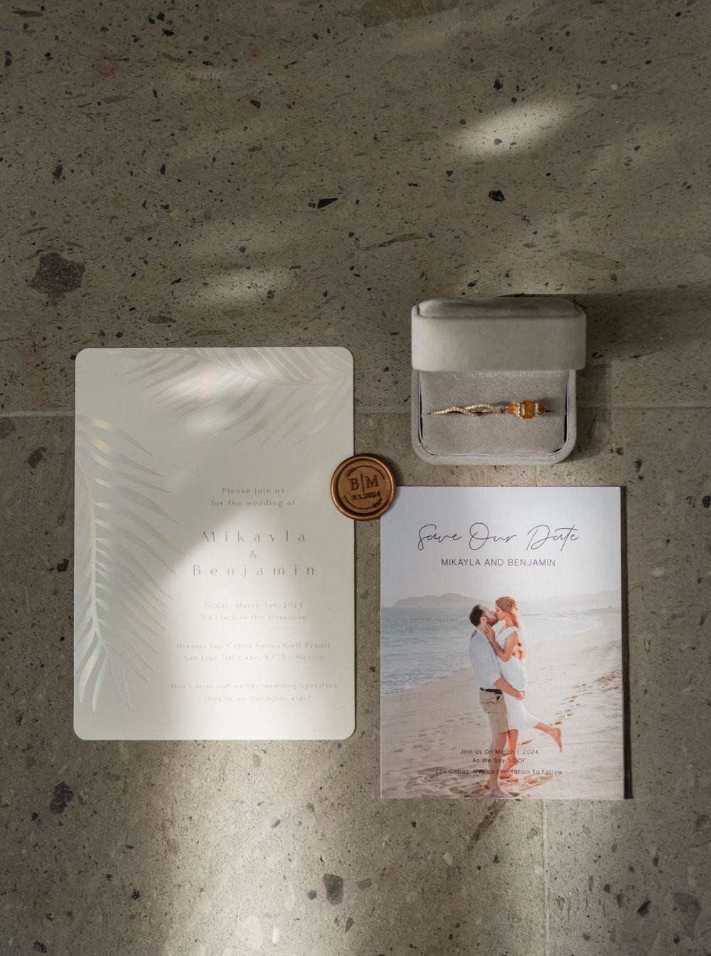 Wedding details flatlay with invitation, ring box, and wax seal on concrete background.