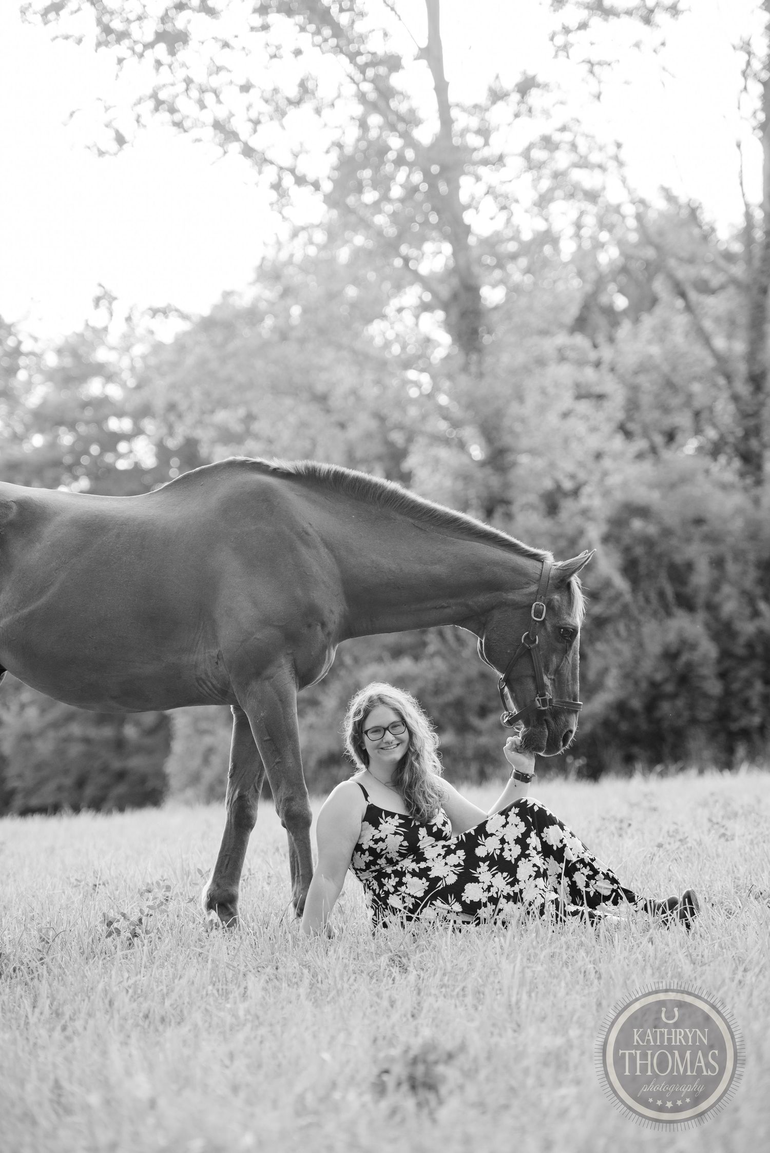 A black and white portrait of a horse and someone in a patterned dress sitting in a field.