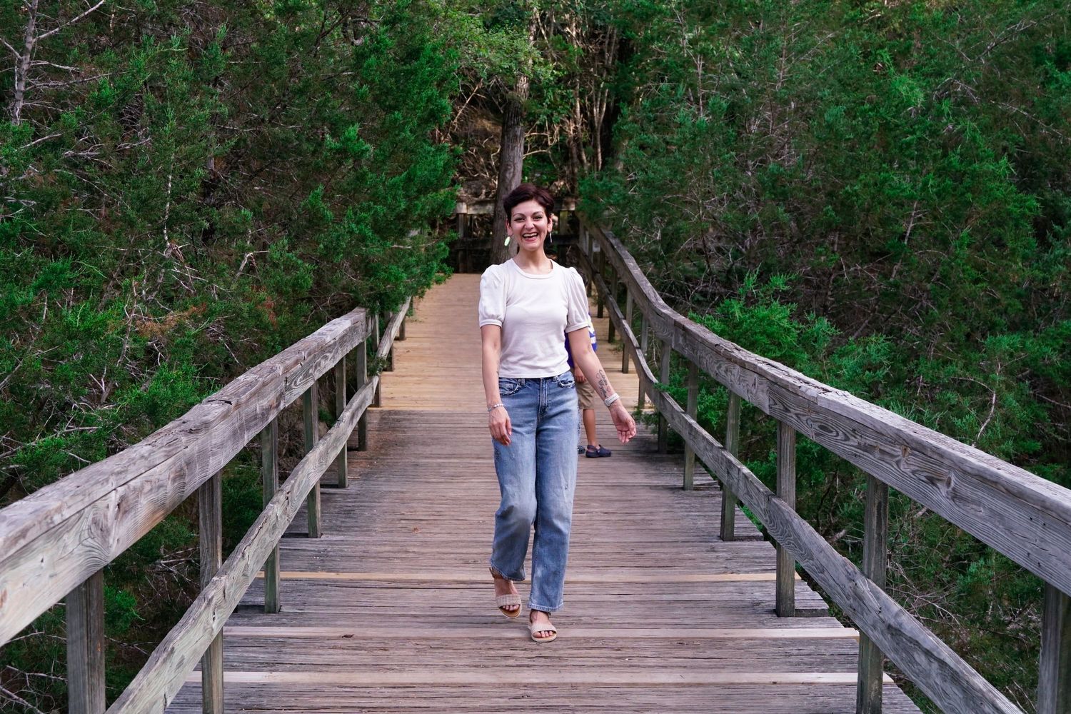 A person in jeans and white shirt walks barefoot across a wooden boardwalk surrounded by greenery.