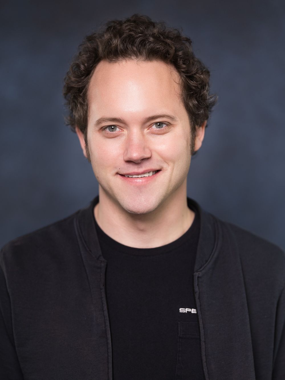 Professional headshot portrait of a person in a black jacket against a dark background.