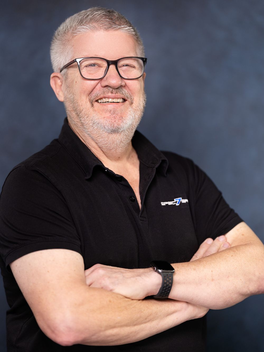 Professional headshot of a person wearing glasses and a black polo shirt, smiling warmly with arms crossed.