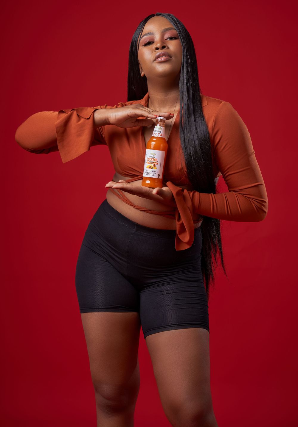 Fashion photography with model in brown top holding juice bottle against red backdrop.