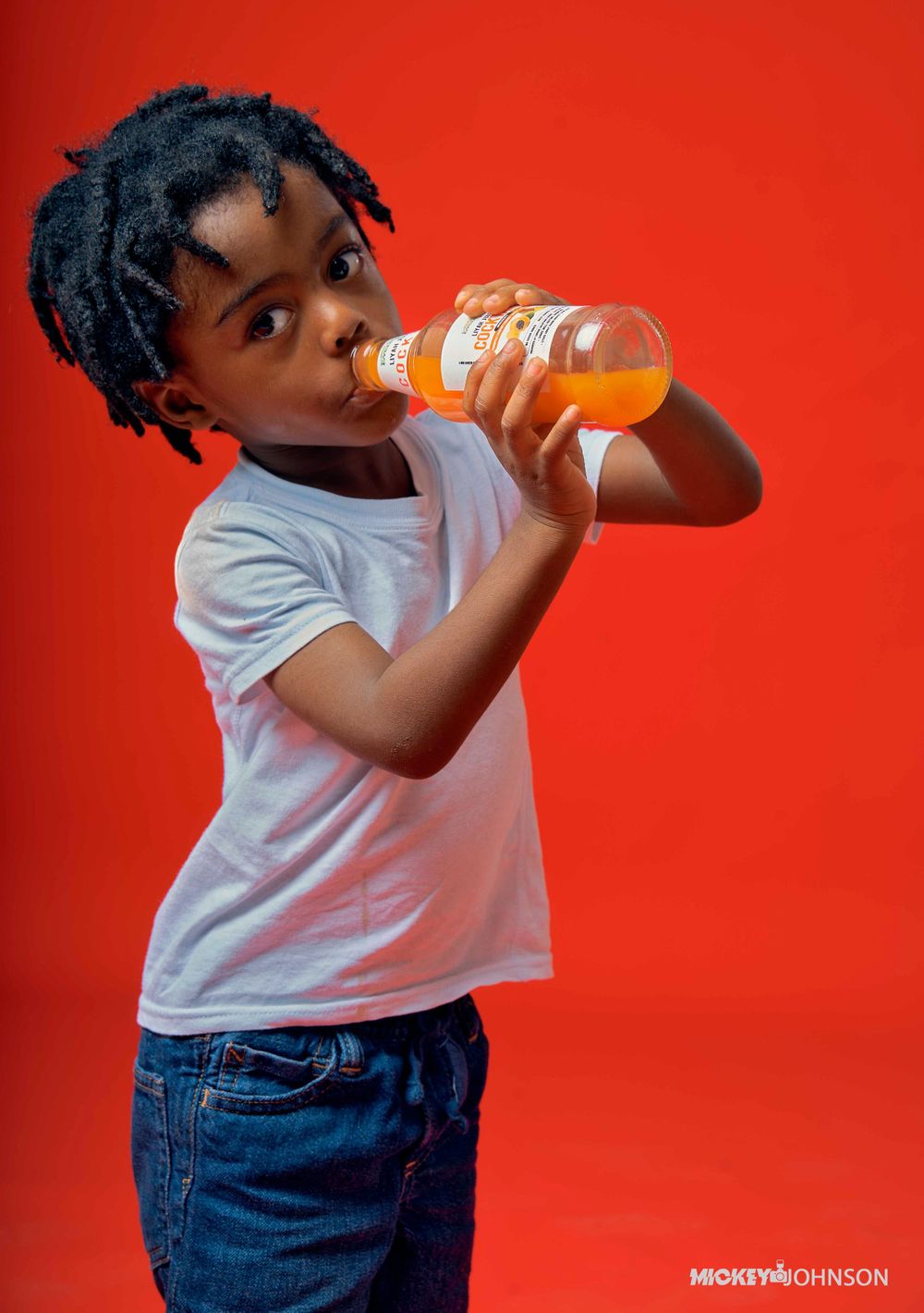 Child in white t-shirt and jeans drinking from an orange juice bottle against red background.
