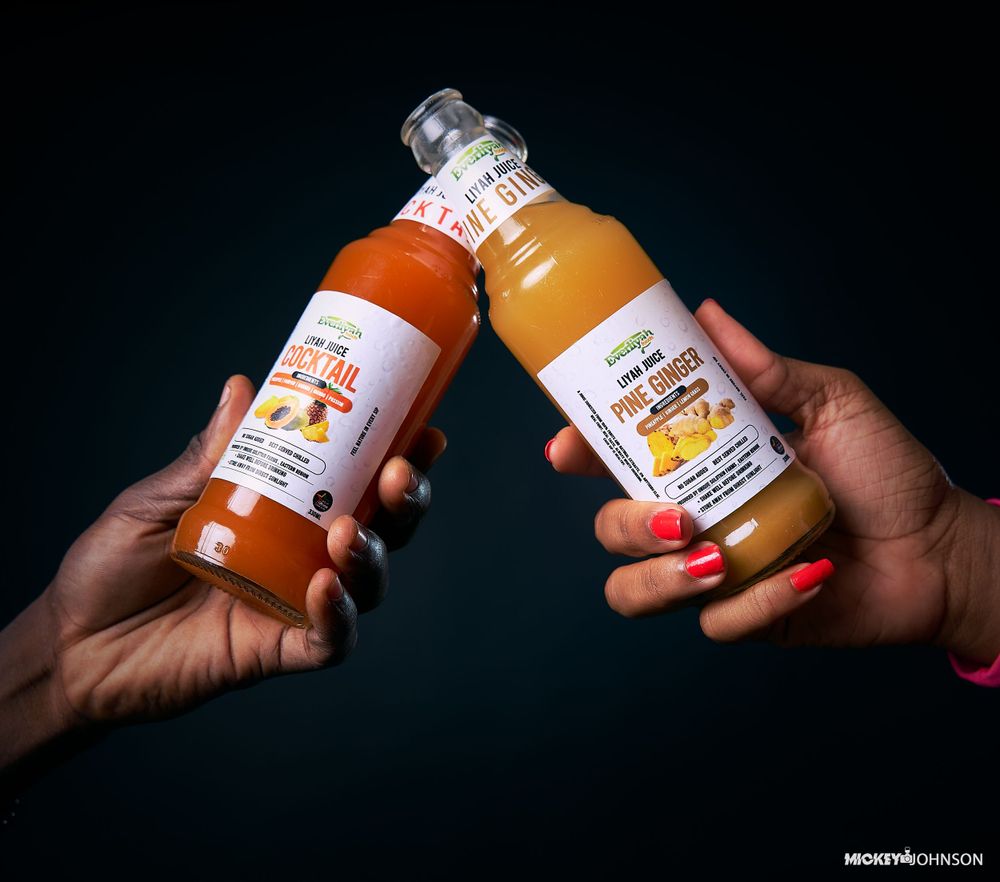 Two hands holding orange juice bottles against a dark background in a product photography setup.