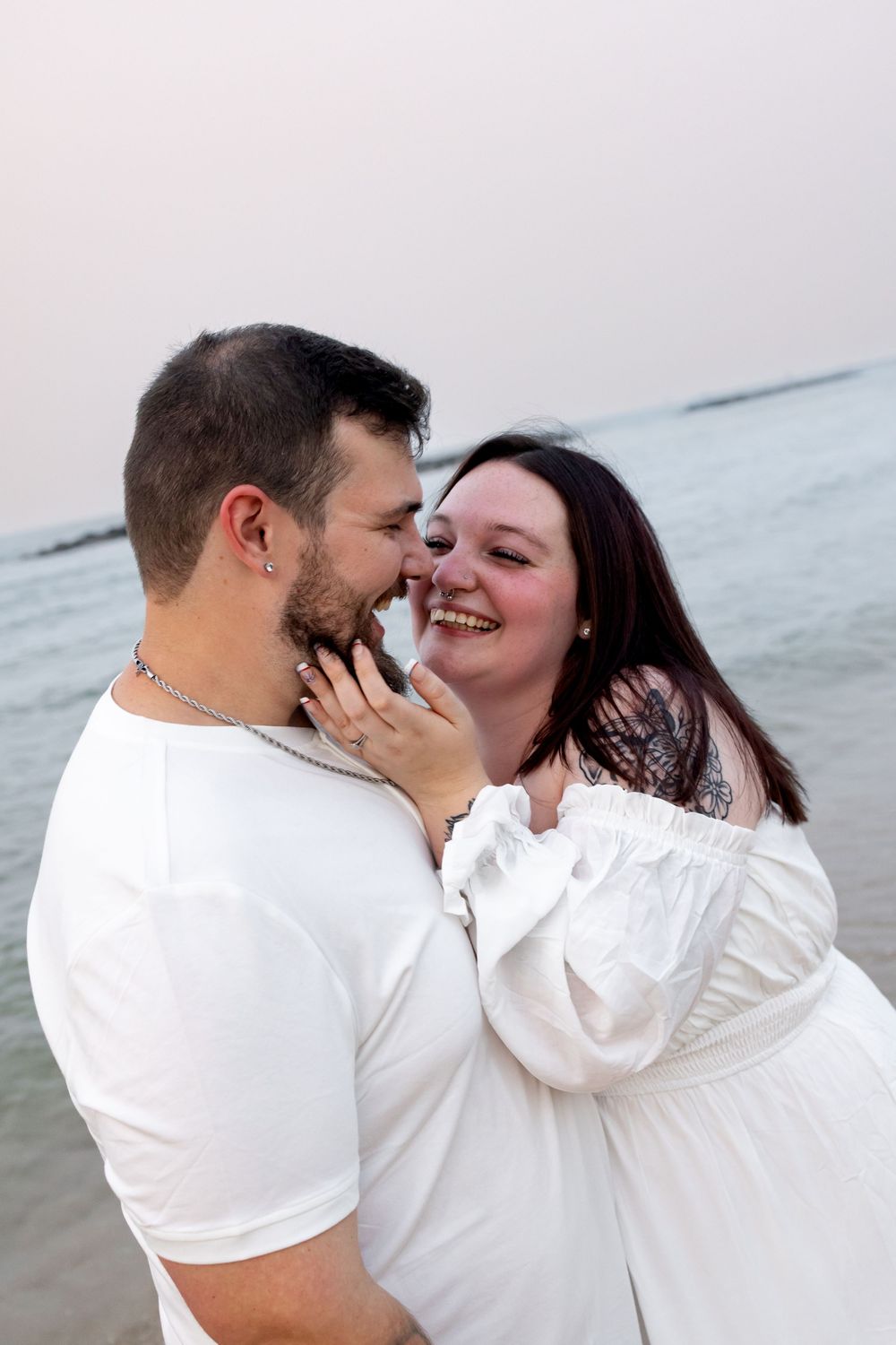 Happy couple in white outfits sharing a romantic moment on the beach, smiling and embracing near the ocean.