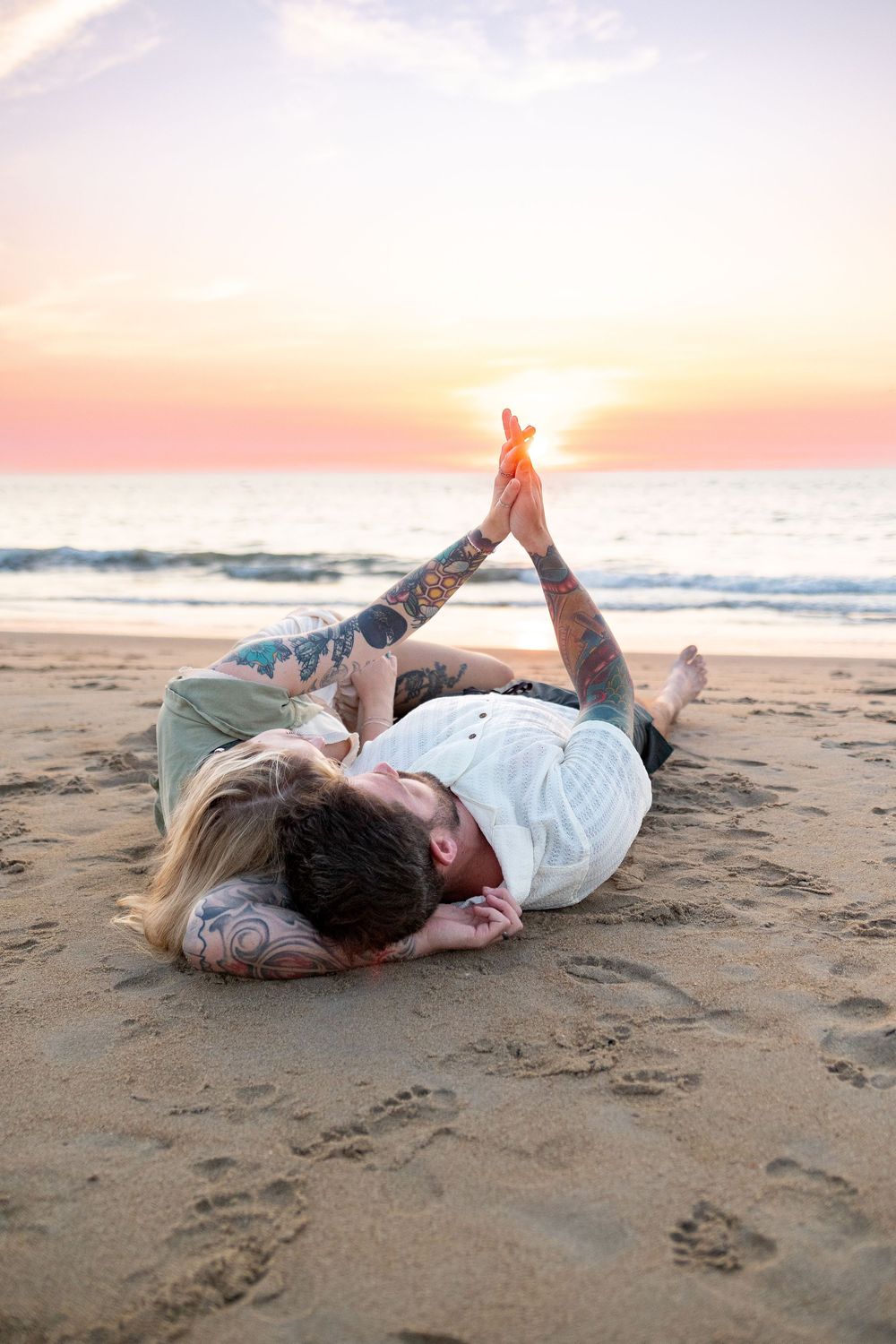 Two people lying on a sandy beach at sunset creating heart shapes with their hands against the golden sky.
