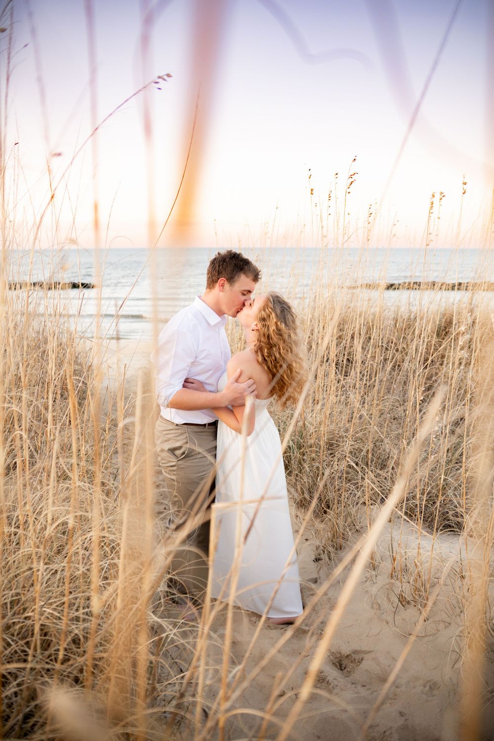 A romantic couple embraces among tall beach grass at sunset by the ocean.