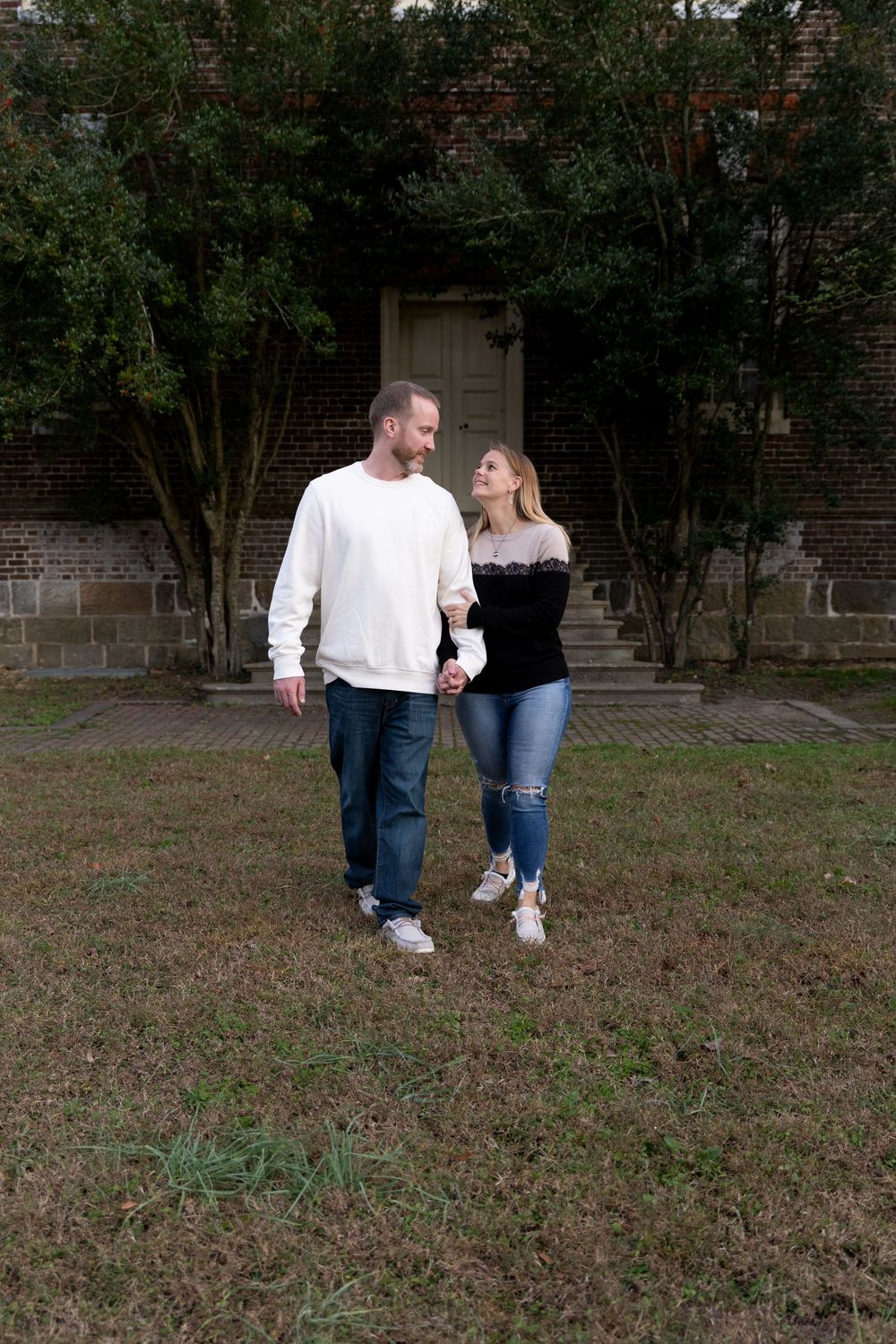 A couple takes a stroll together near a brick building surrounded by greenery and trees.