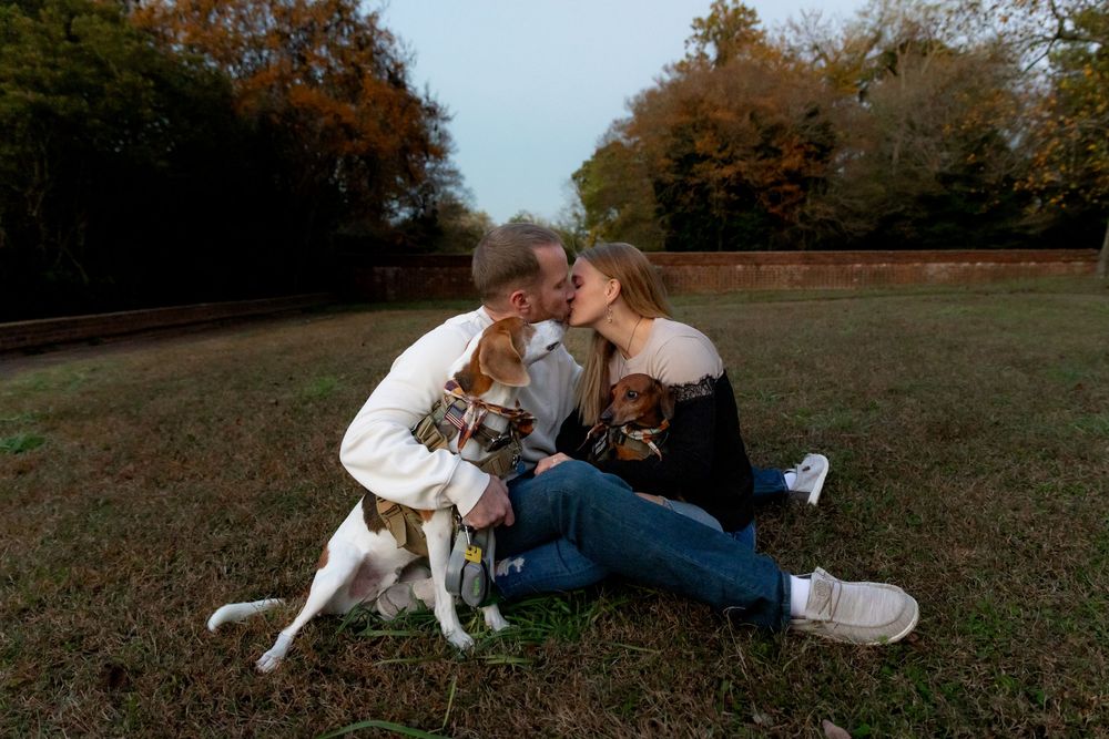 A couple sits on grass in autumn with their small white dog, sharing an intimate moment together in a park.