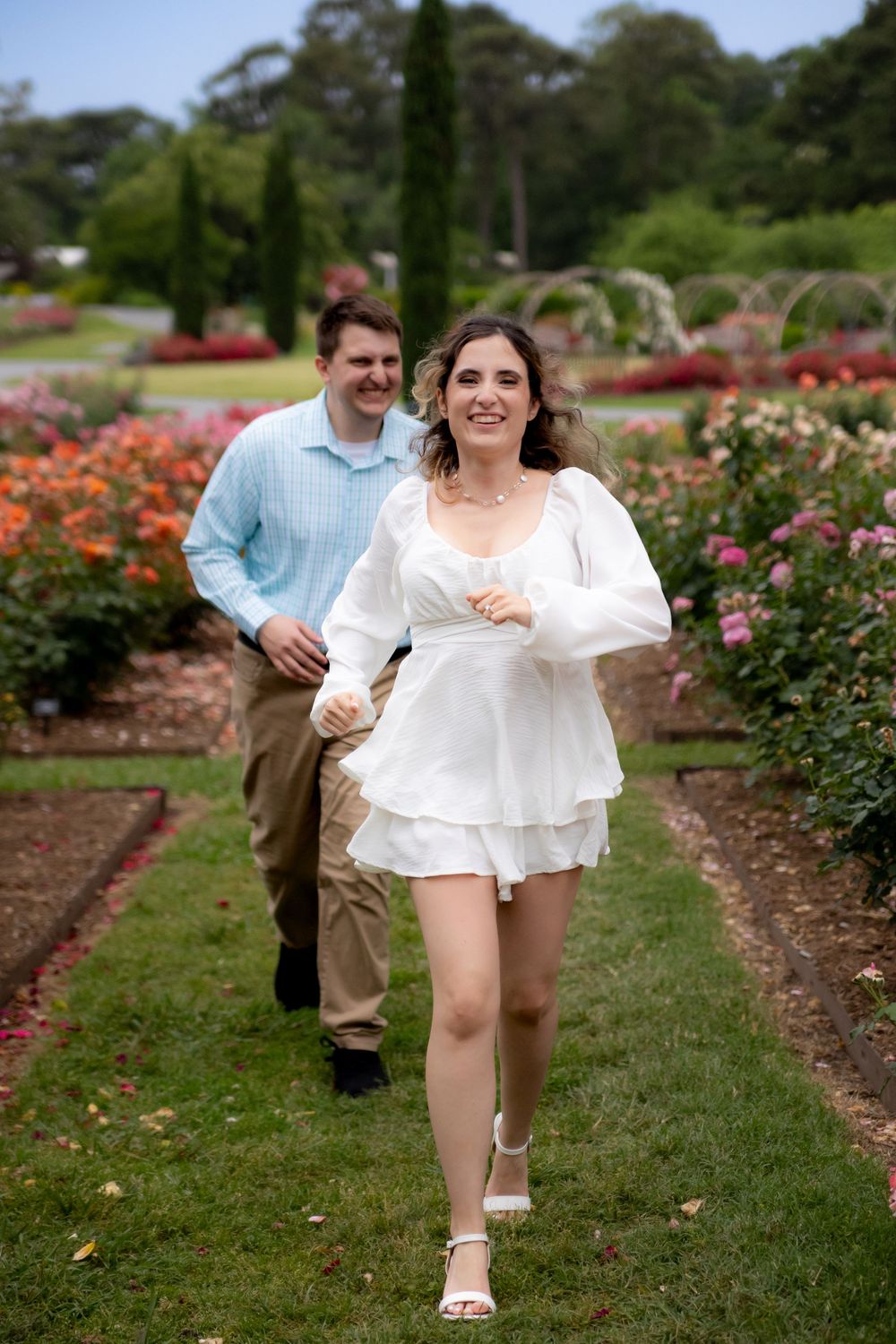 A couple walking together through a lush garden path lined with blooming roses and tall cypress trees.