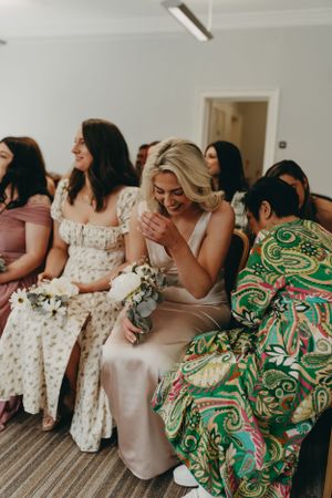 A group of wedding guests in colorful dresses laugh together while seated on a bench. Wedding photographer, Devon & South Hams